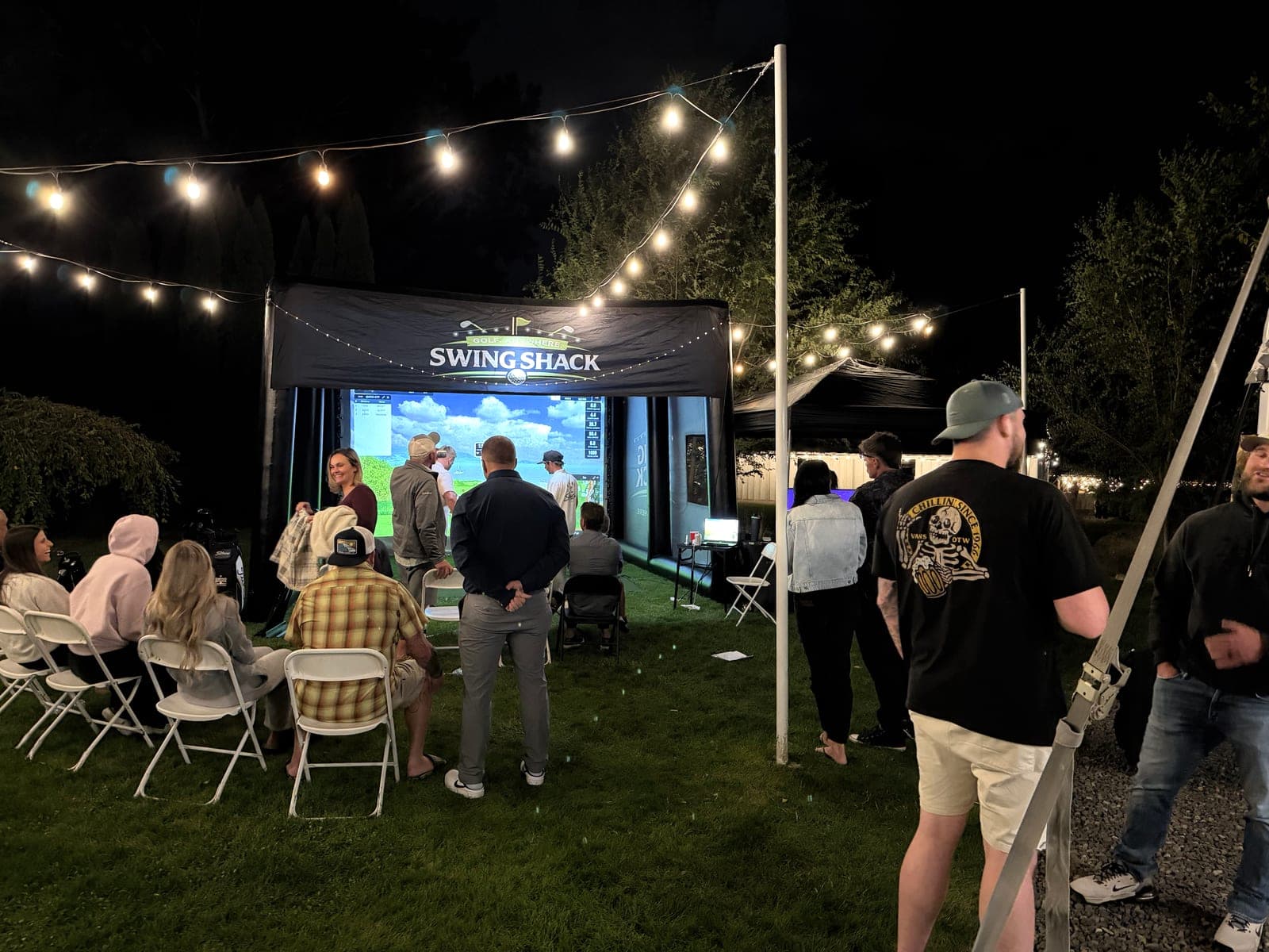 Guests gathered on a lawn under string lights around the glowing Swing Shack simulator at a nighttime backyard party
