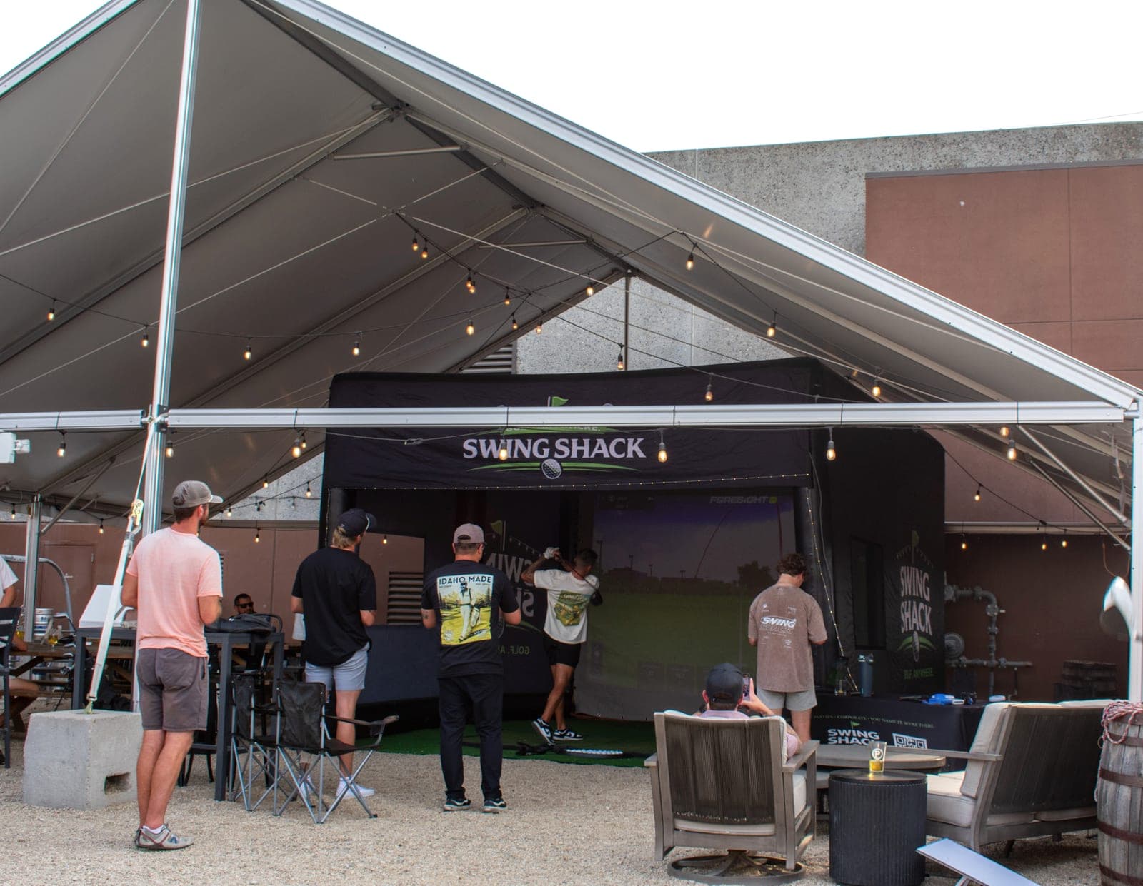 A golfer swinging in the Swing Shack simulator under a large tent at an outdoor venue while guests watch